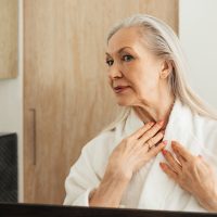 A mature woman in a bathrobe looking at her neck wondering how to get rid of her turkey neck, neck cords, and neck lines