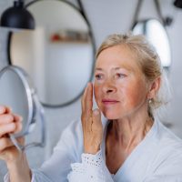 A photo of a woman looking in the mirror at her cheeks and considering broken capillaries and Rosacea Treatment